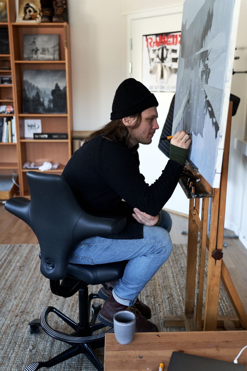 Artist painting at an easel in side profile, sitting forward on the saddle seat of a black leather HÅG Capisco 8106 with a black footring and five-star base.