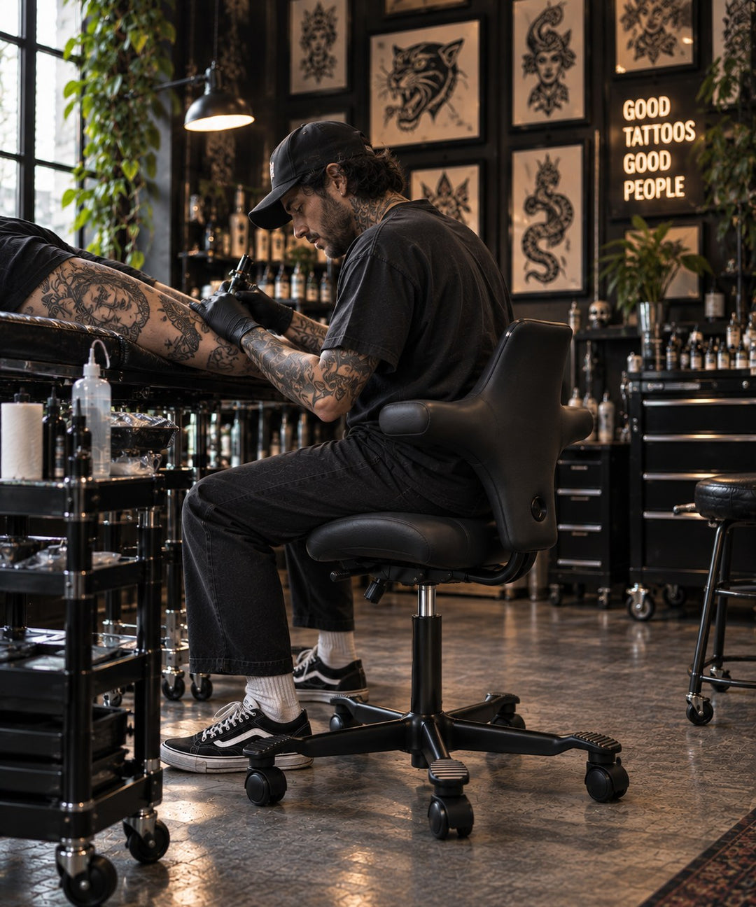 Tattoo artist in a black cap leaning forward on a black leather HÅG Capisco 8106 to work on a client's forearm, in a studio hung with framed flash art.