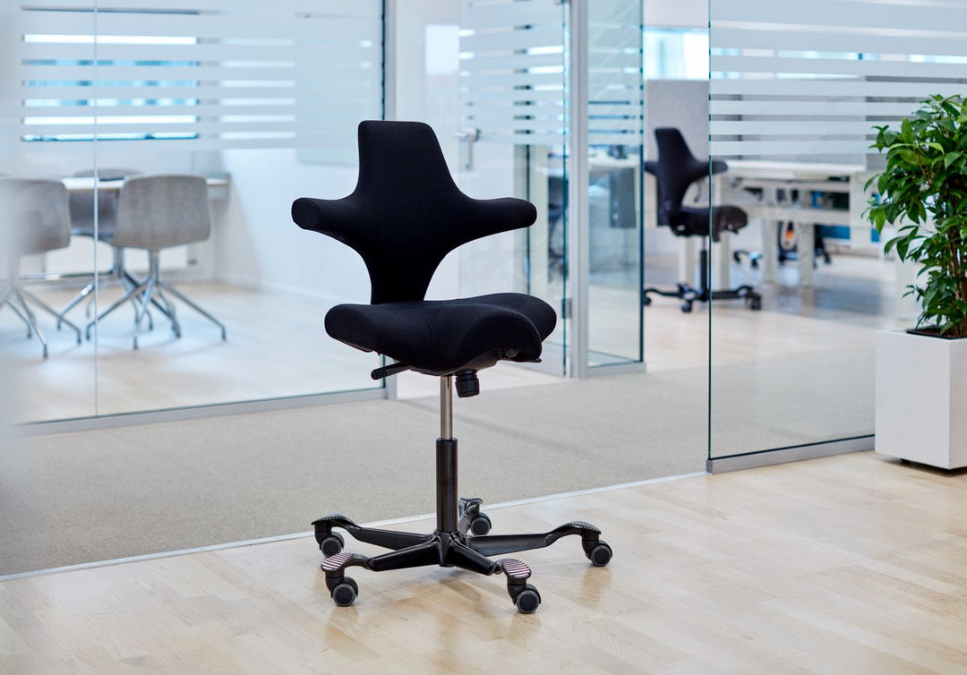 Single black fabric HÅG Capisco 8106 with a black gas lift and five-star base standing on a pale wood floor beside glass partition walls in a modern office.