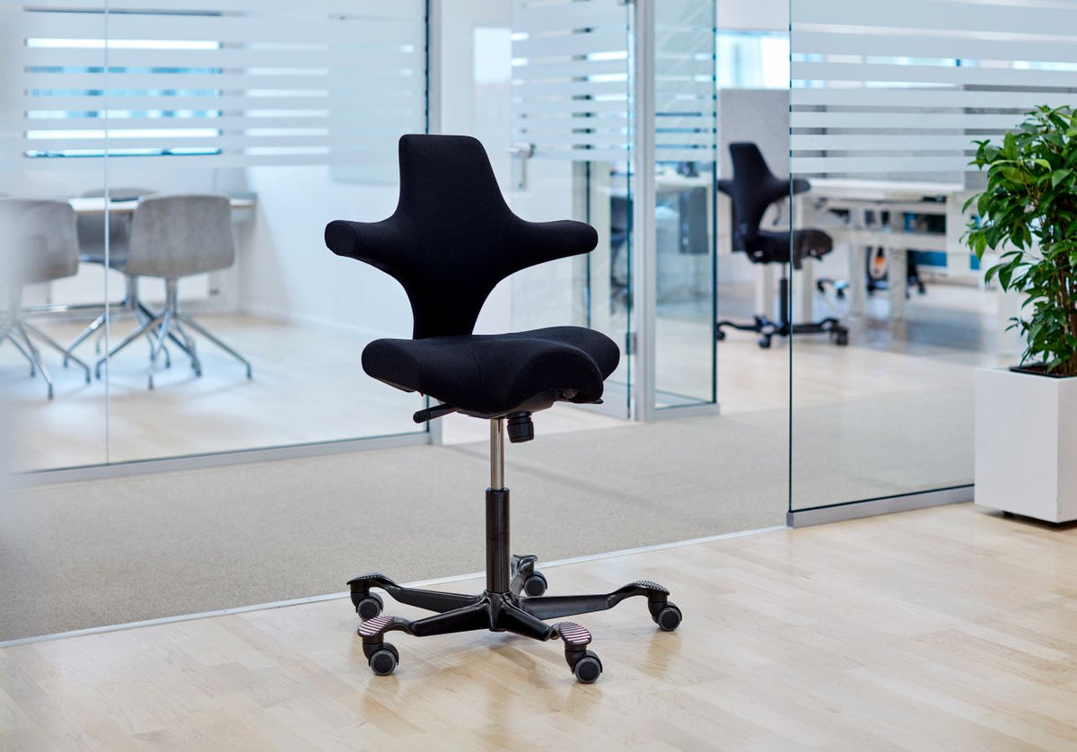 Single black fabric HÅG Capisco 8106 with a black gas lift and five-star base standing on a pale wood floor beside glass partition walls in a modern office.