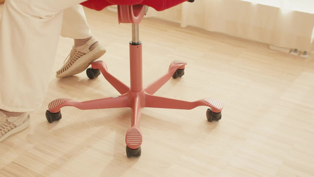 Detail of the coral-pink five-star base, castors and gas lift cylinder of a HÅG Capisco on a pale wood floor, with a seated person's feet beside it