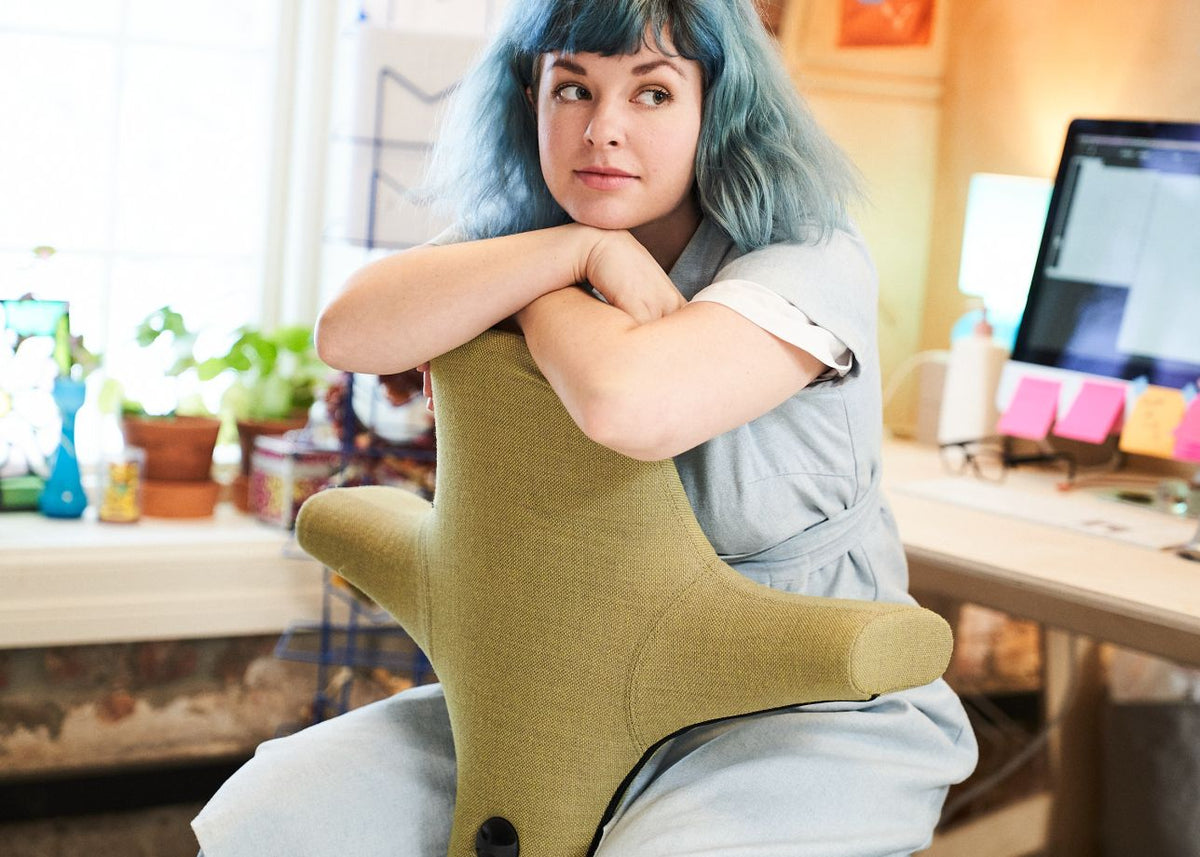 Woman with blue hair sitting astride an olive-green HÅG Capisco 8106, her folded arms resting on top of the winged backrest, with a desk and monitor behind her.