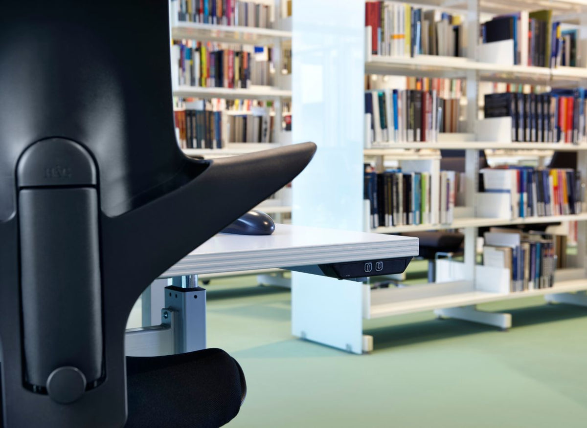 Close-up of the black backrest and its HÅG-branded height-adjustment lever on a Capisco pulled up to a height-adjustable desk in a library of full bookshelves.