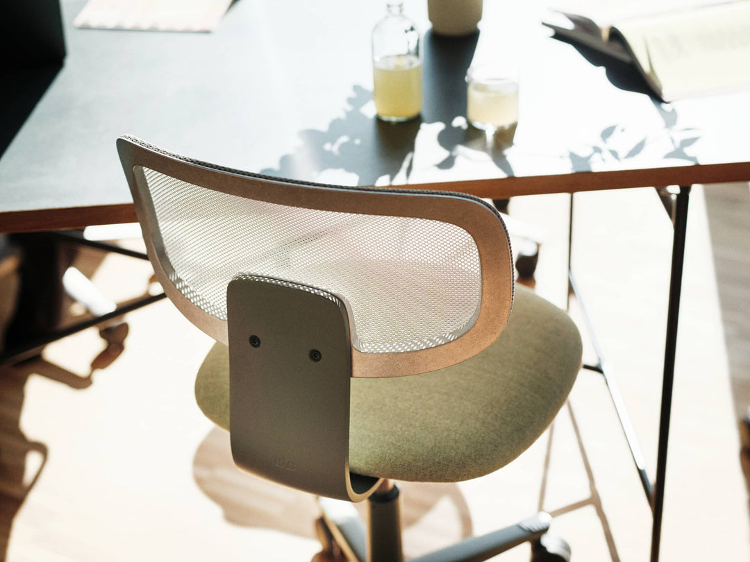 Close-up from above of a HÅG Tion with a white mesh backrest in a beige frame and olive-green seat, tucked under a sunlit pale grey desk.