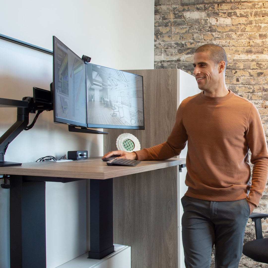 Man standing at a sit-stand desk using screens on a matte black Ergotron HX Dual Monitor Arm