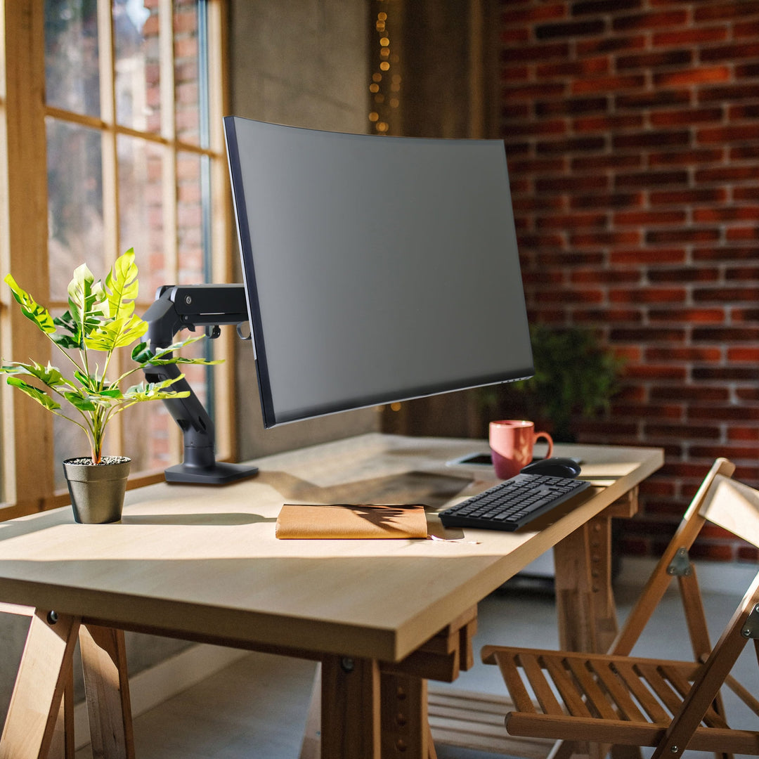 Home office desk by a brick wall with a large curved screen on a matte black Ergotron HX Desk Monitor Arm