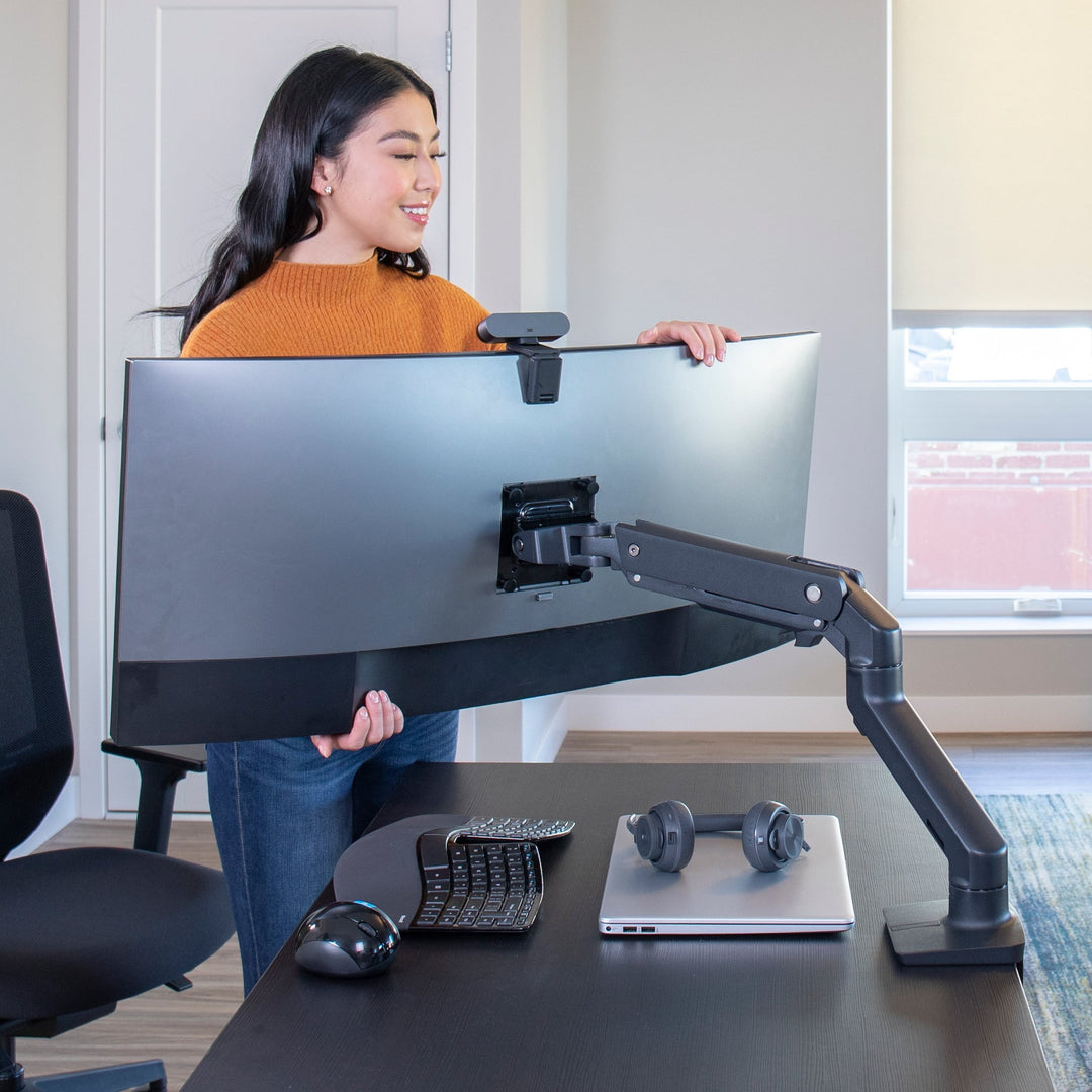 Woman lifting a large curved screen onto a matte black Ergotron HX Desk Monitor Arm