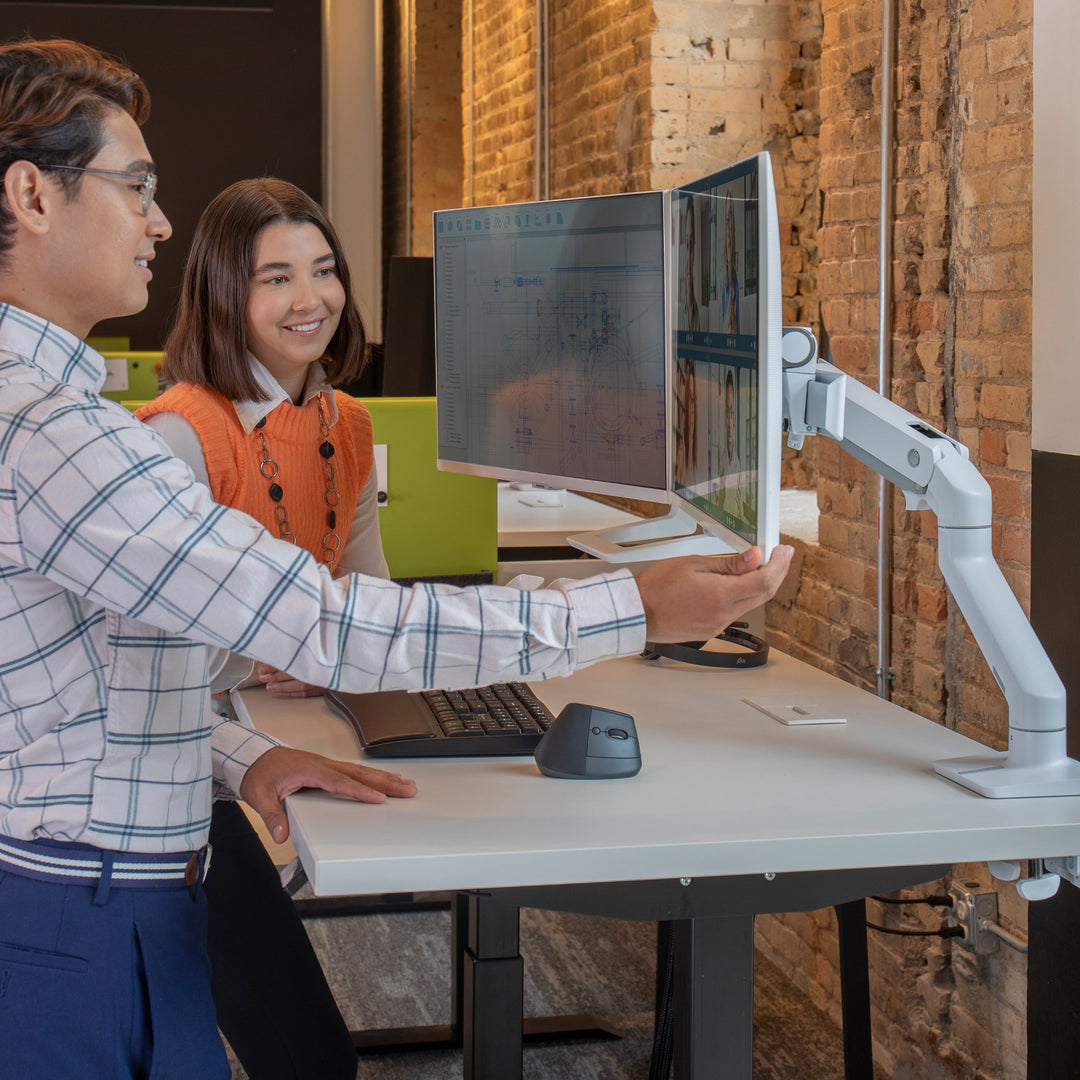 Two people adjusting a screen on a white Ergotron HX monitor arm at a standing desk