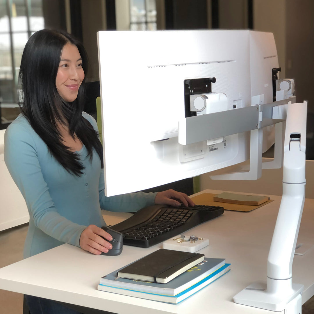 Woman working at a standing desk with a large screen on a white Ergotron HX monitor arm