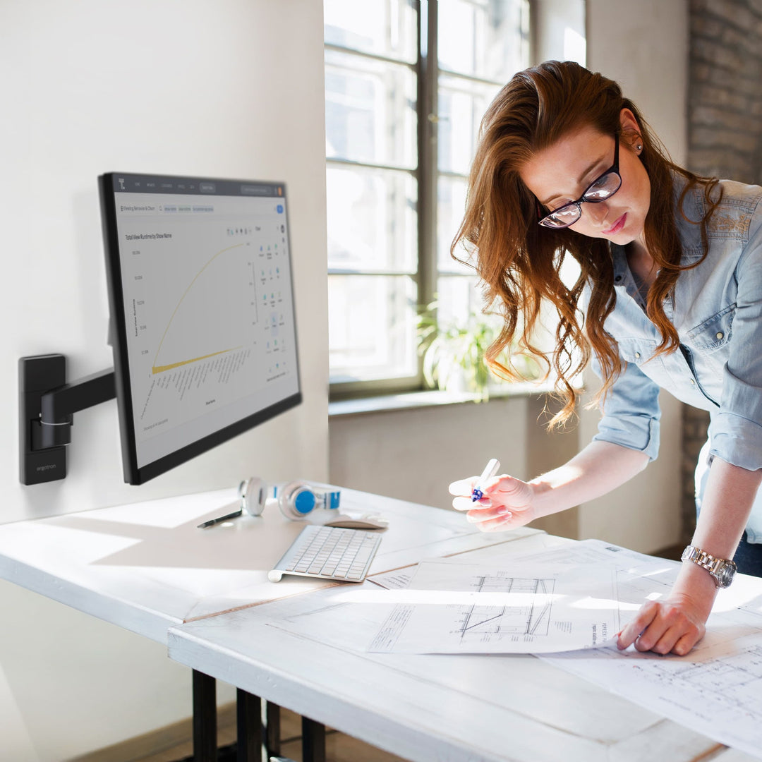 Woman working at a drafting table beside a screen on a matte black Ergotron LX Pro Wall Monitor Arm