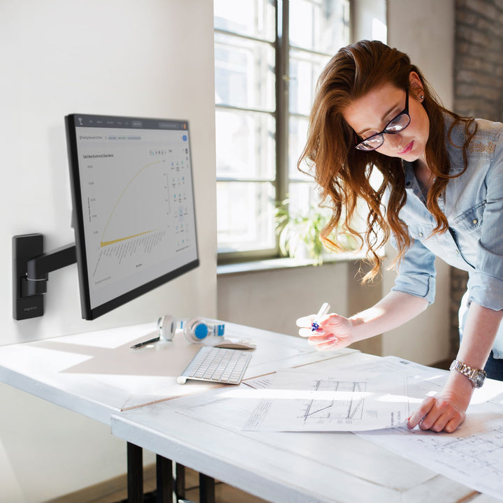 Woman working at a drafting table beside a screen on a matte black Ergotron LX Pro Wall Monitor Arm