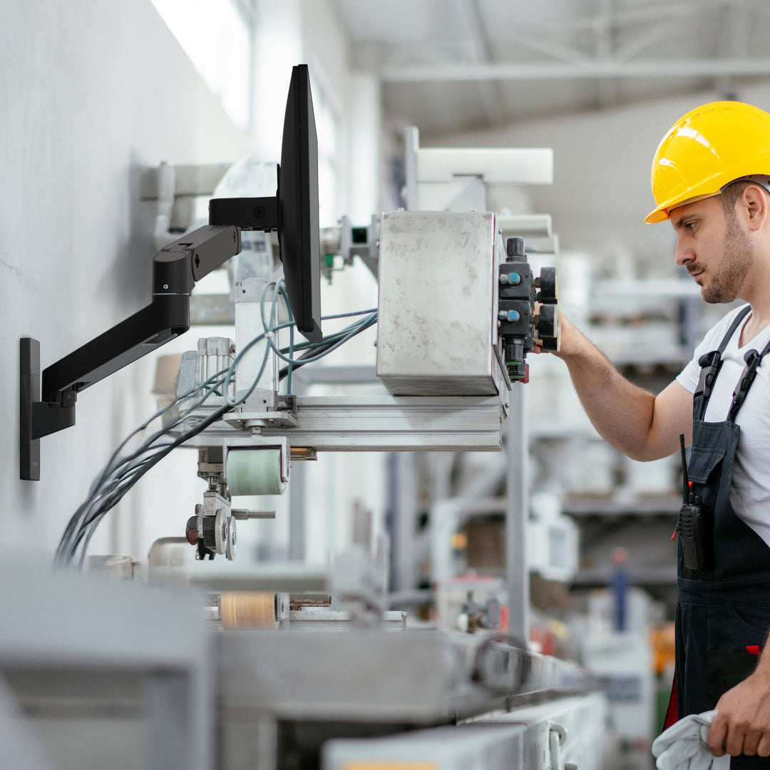 Matte black Ergotron LX Pro Wall Monitor Arm holding a screen beside machinery, with a worker in a hard hat