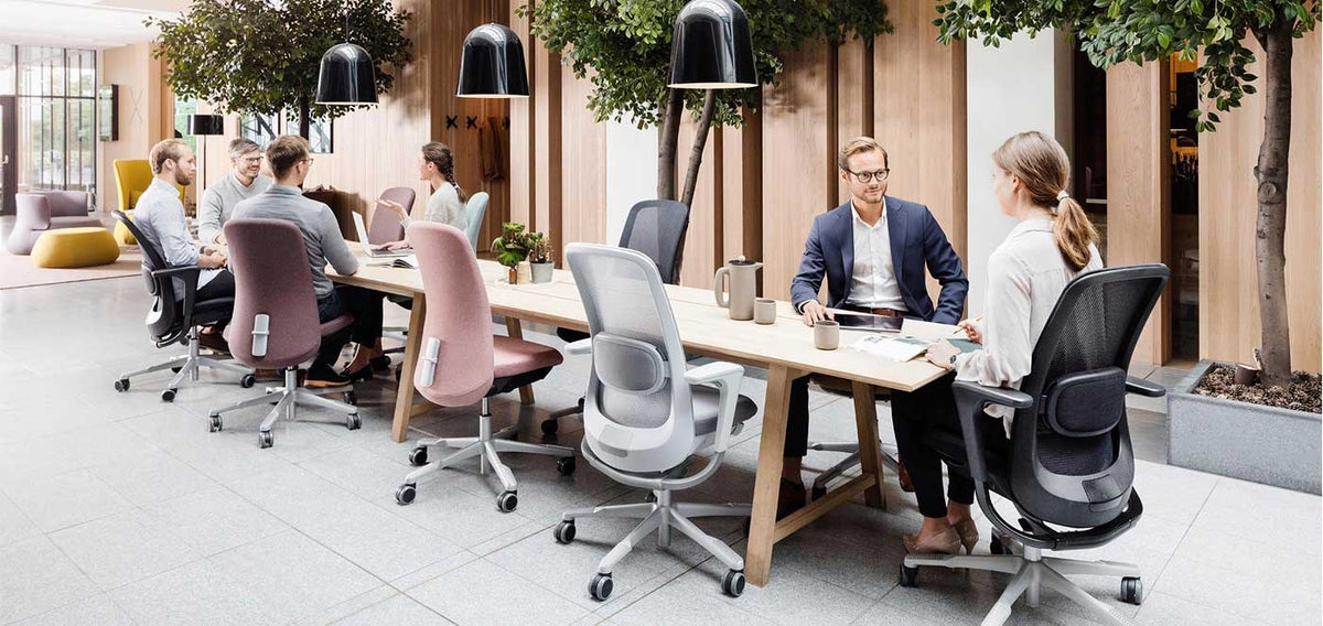Man in a navy blazer talks with a woman seated in a dark HÅG SoFi mesh chair at a long timber table, pink and grey SoFi chairs nearby. - Chair Dinkum