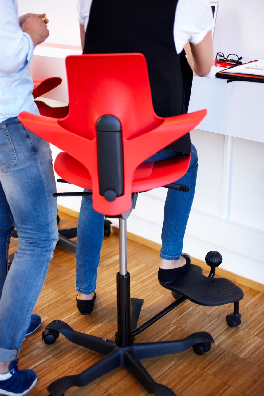 Person perched on a red HÅG Capisco Puls at a sit-stand desk, one foot resting on the plate of a black HÅG StepUp beside the ball