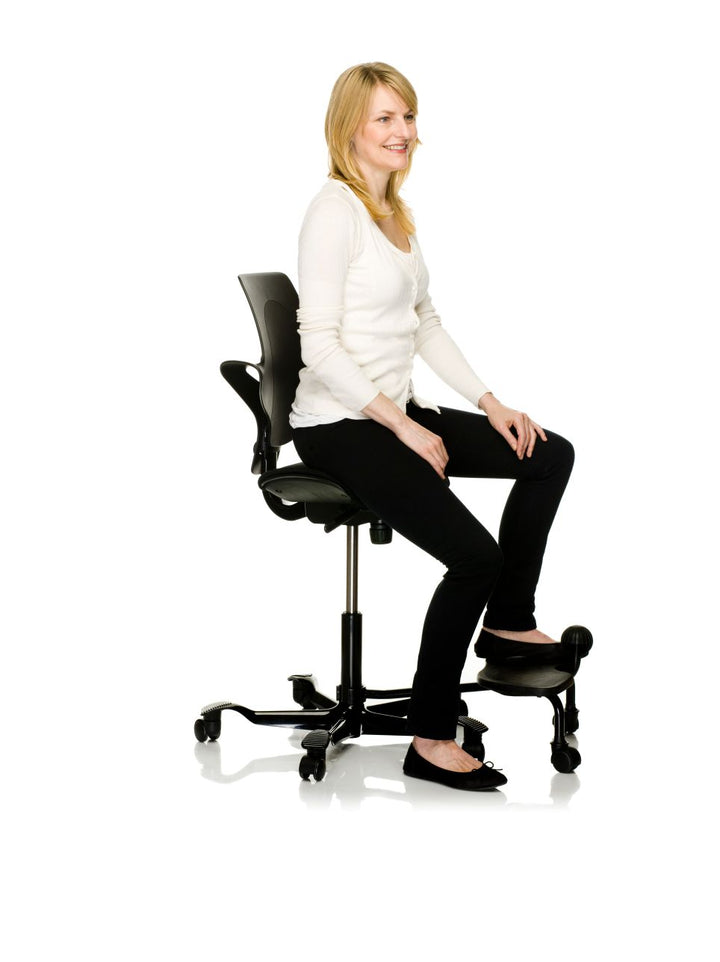 Woman seated on a black HÅG Capisco Puls with one foot on the floor and the other on a black HÅG StepUp