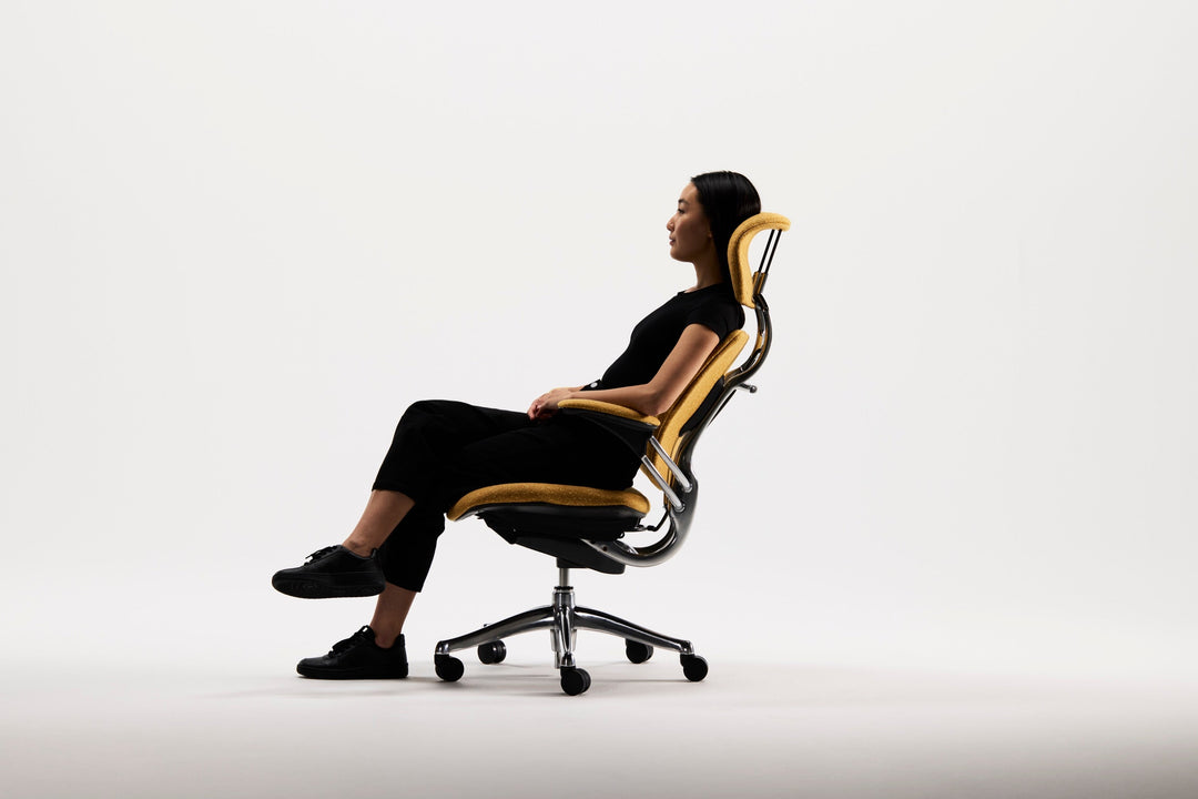 Woman reclining in a yellow Humanscale Freedom chair with polished aluminium trim, side view in a studio - Chair Dinkum Australia