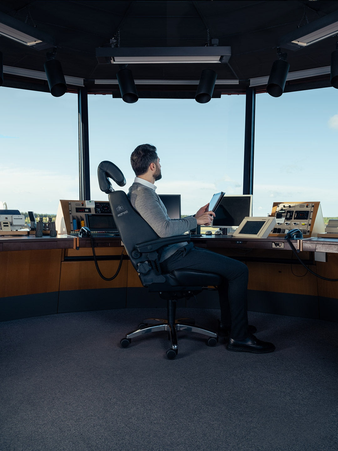 Operator seated in a black RH Secur24 in a control tower, reading from a tablet with the runway behind - Chair Dinkum Australia